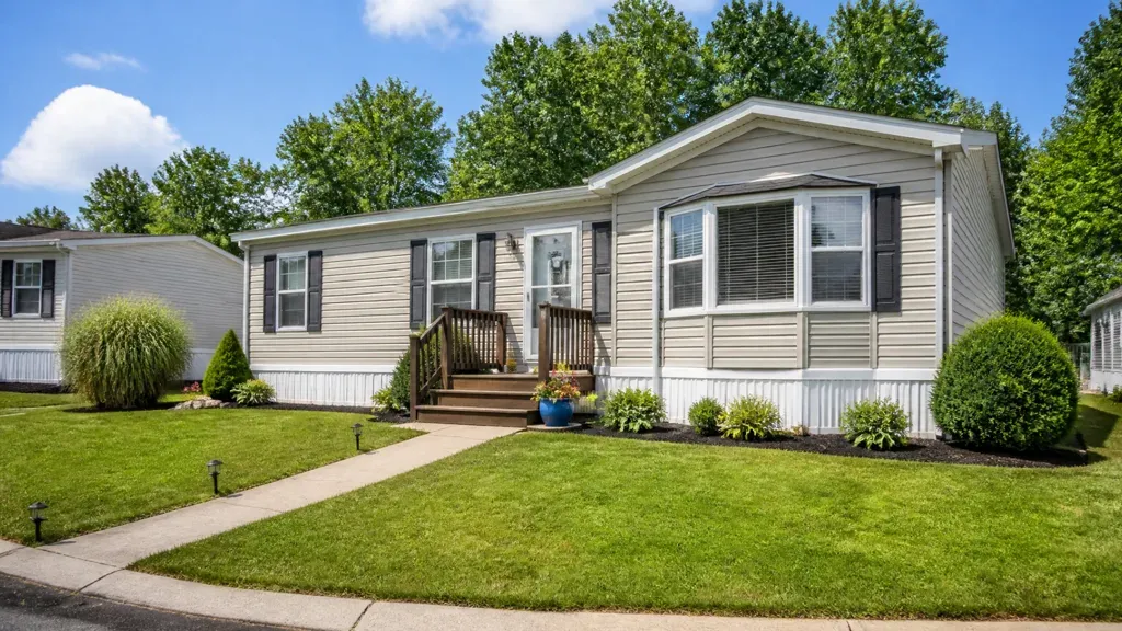A grey mobile home with a bay window, wooden steps and a neatly kept front lawn.